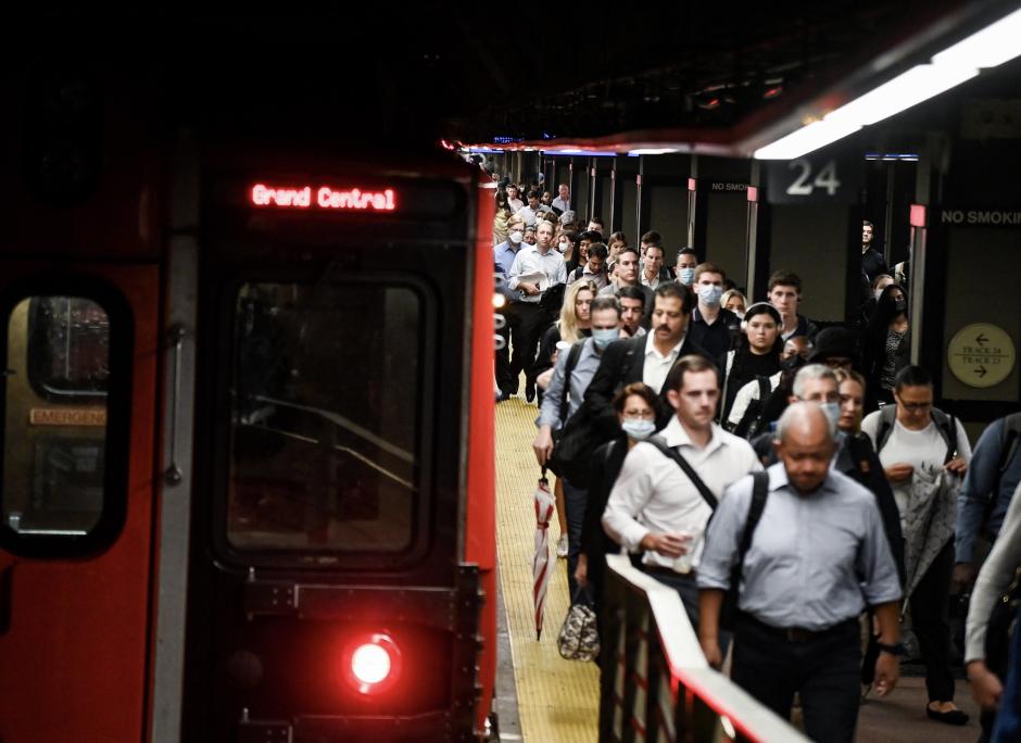 A group of people walks on a train platform. They are walking next to a red train with an electronic display that says "Grand Central."