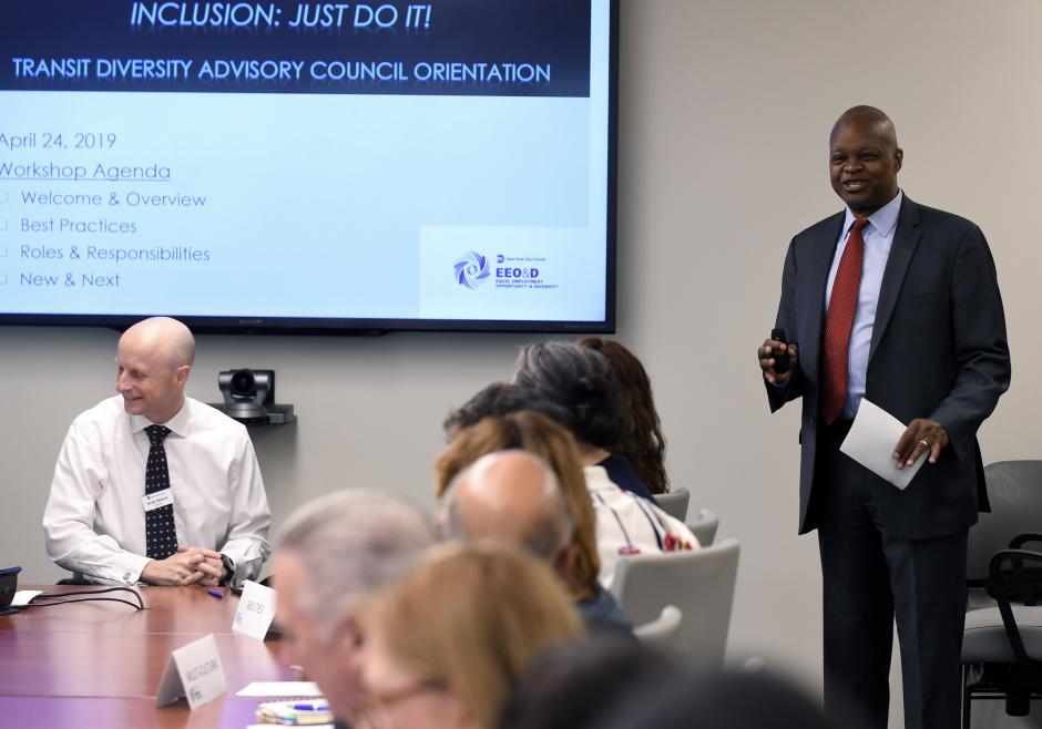 A man smiles as he speaks to a group of people at a conference room table. A presentation slide is visible on a projection screen. It reads "Inclusion: Just do it!" and "Transit Diversity Advisory Council orientation."